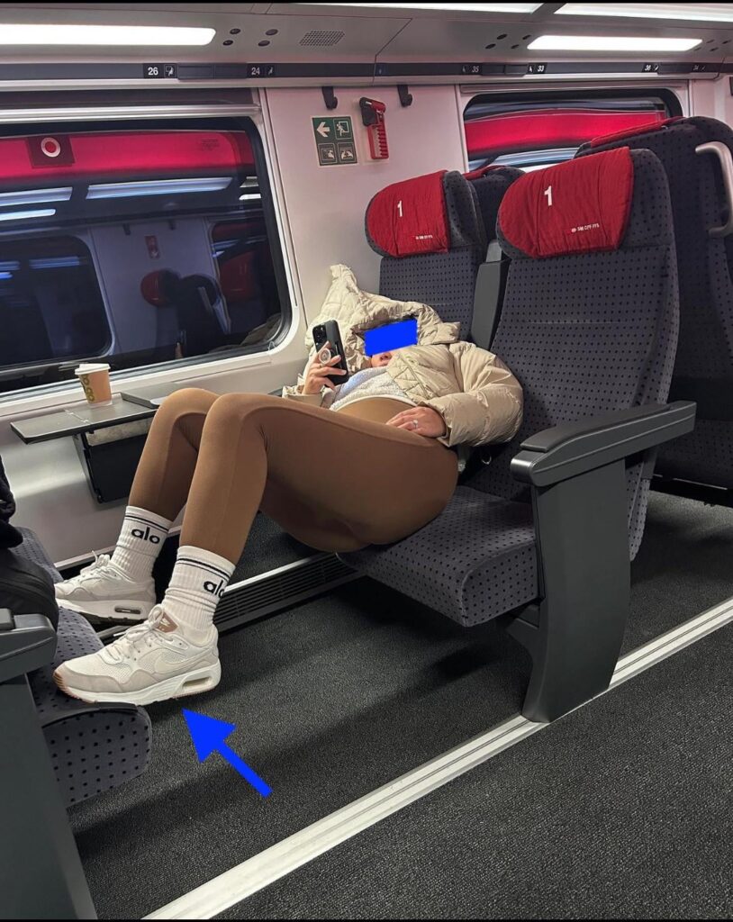 スイスでファーストクラスに乗っている米国人旅行者が、携帯電話で破滅的なスクロールをしながら、何気なくスニーカーを座席に置いている...