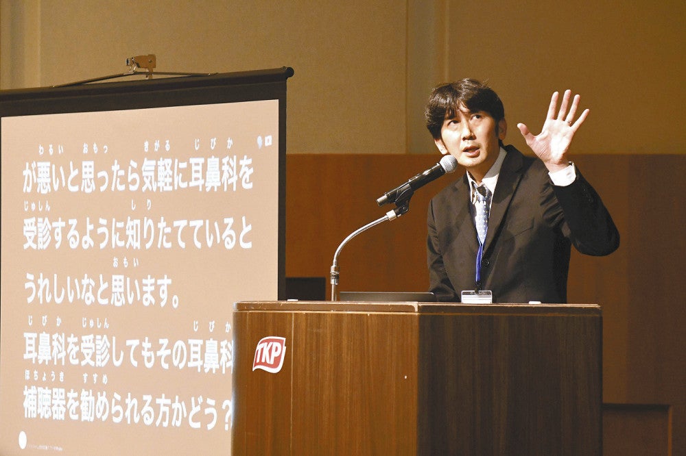 フォーラムで「気軽に補聴器を使ってほしい」と話す野田さん。会場では耳が遠い人のために発言の字幕が映し出された（福岡市で）