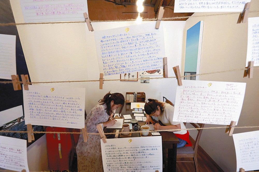 自分宛ての手紙がつるされた店内で「１年後の私」に向けて筆を走らせる戸田さん（左）。さまざまな思いがあふれるこの場所で自分と向き合う真剣な時が流れる