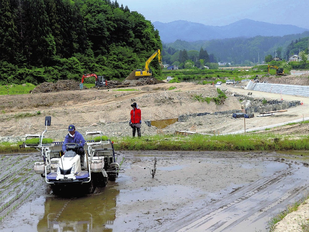 豪雨災害で被災した水田で、２年ぶりに田植えをする佐原さん。後方では川の改修工事が進む（２４日、飯豊町小白川で）