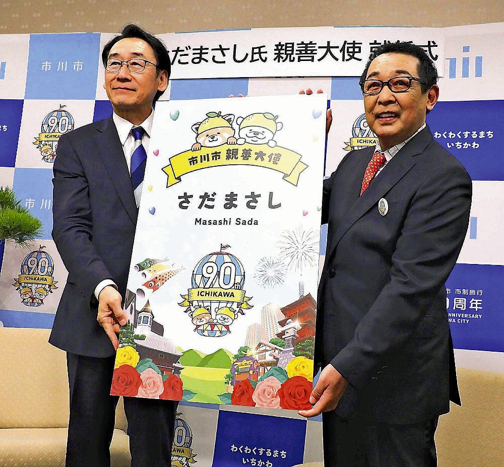 親善大使の名刺の拡大版を手にするさださん（右）（２４日、千葉県市川市文化会館で）