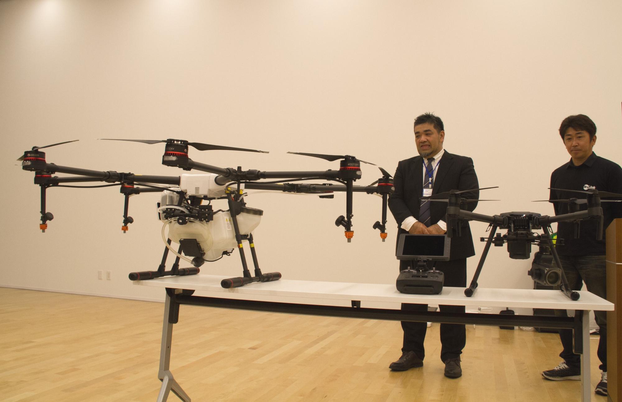 2機の無人航空機が飛んでいる様子を後方で2名の男性が見ている写真