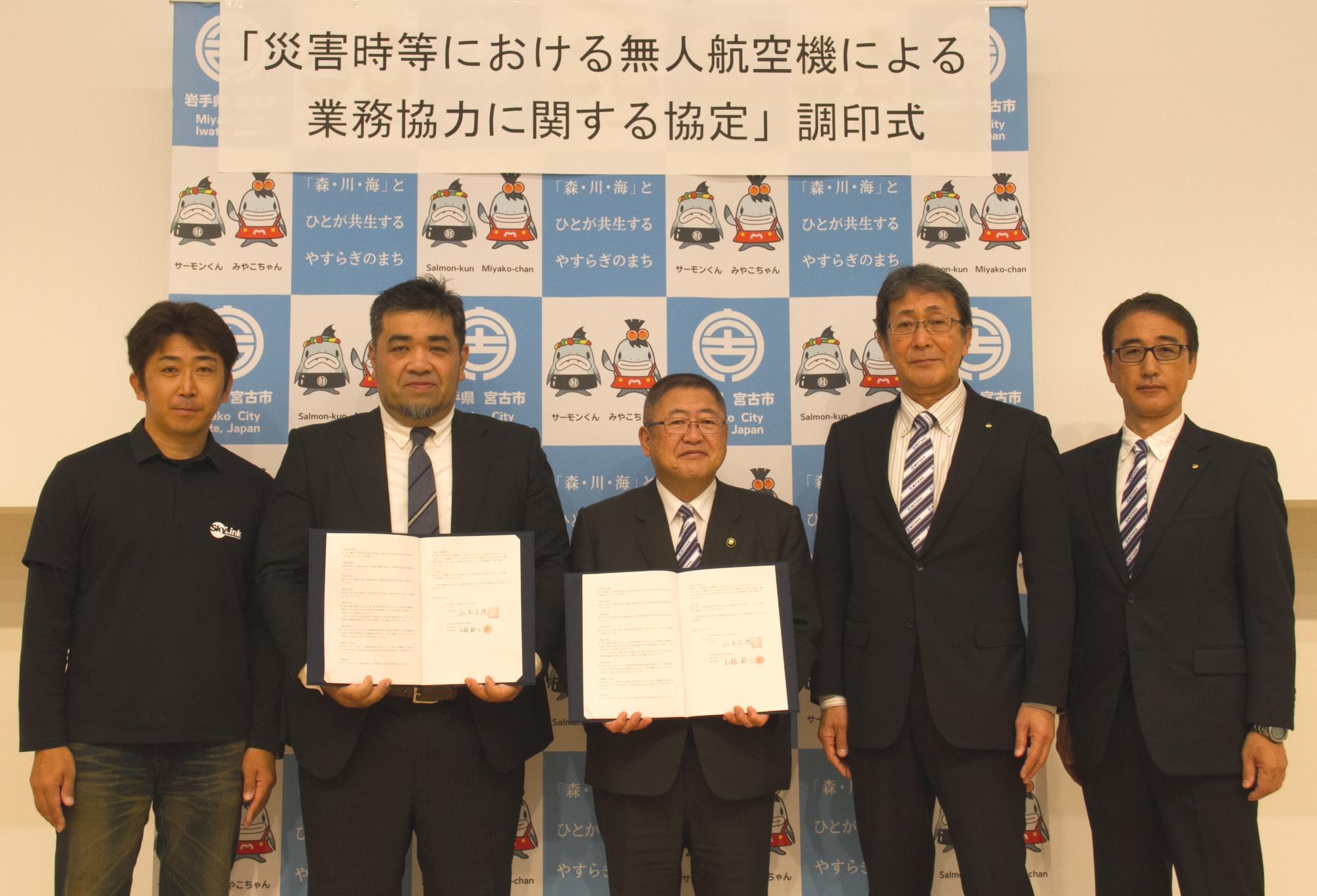 5名の男性が1列に並び左から2番目と3番目の男性が協定書をも持ち記念撮影をしている写真