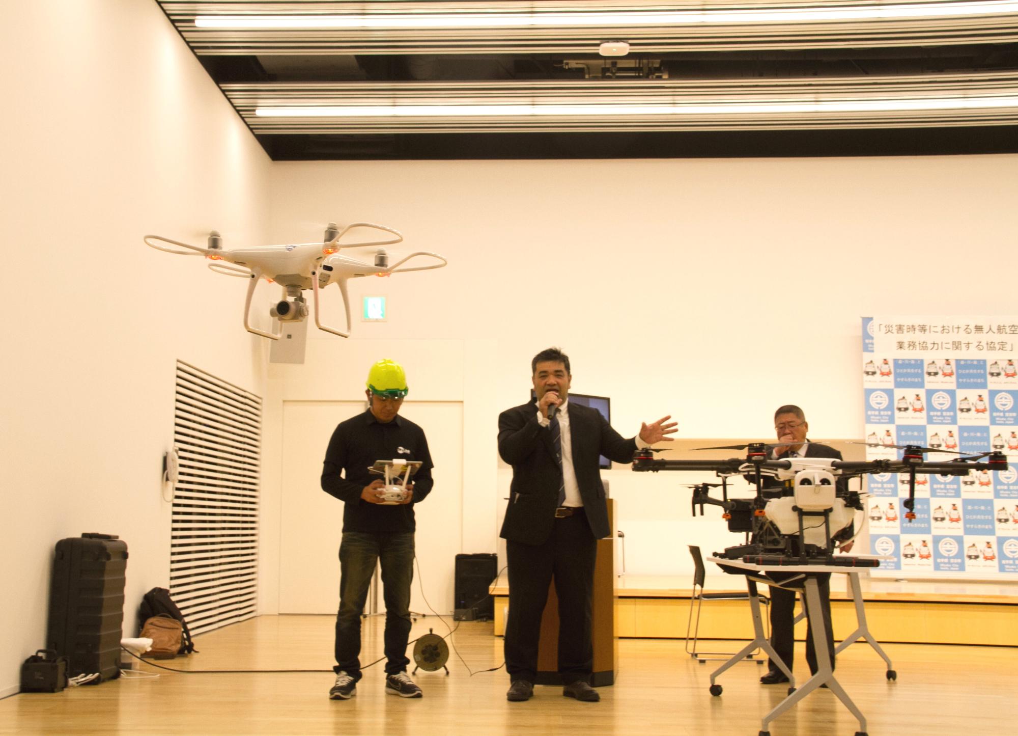 無人航空機の横で桐田教男副市長がマイクで説明をし、1人の男性が操縦をして山本宮古市長は無人航空機の後ろに立っている写真