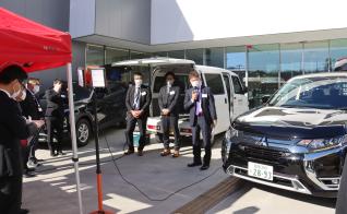 バックドアが開けられた白い車の前で関係者3名が並び、右側の男性がマイクを持ち話をしている写真