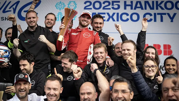 Nissan Formula E driver Oliver Roland in a red race suit surrounded by team members in black shirts celebrating the team's win in Mexico.