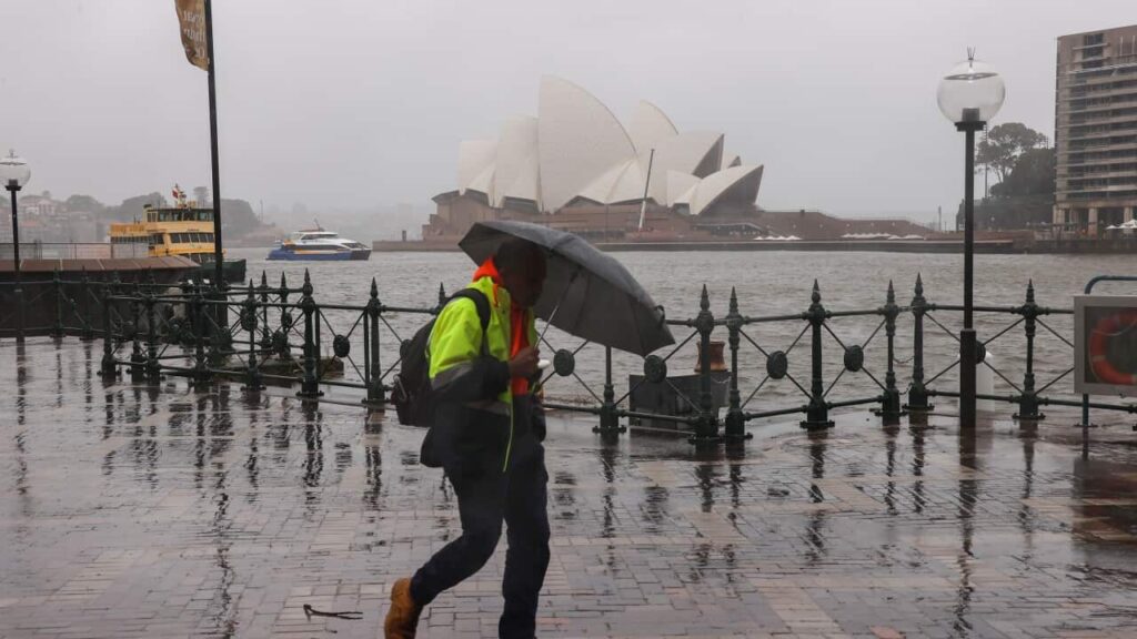 A man wearing a hi-vis jacket, walks in the rain, holding an umbrella, with the Sydney Opera House behind him.