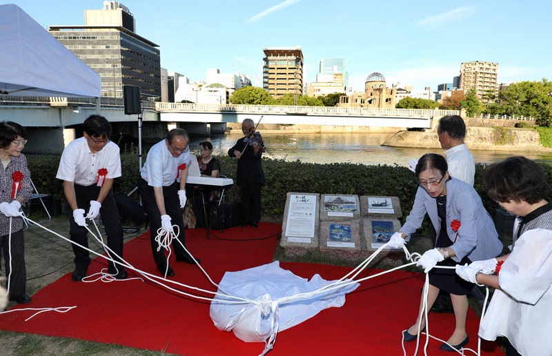 相生橋西詰めの川岸に設置された「原爆の絵碑」の除幕をする関係者。後方は原爆ドーム＝広島市で2025年7月22日午後6時12分、佐藤賢二郎撮影