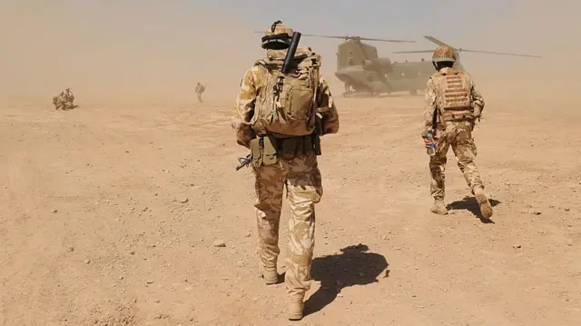 アフガニスタン・ヘルマンド州ナド・エ・アリ地区のピモン軍事キャンプ近くで、軍服を着たイギリス兵が砂ぼこりの舞う砂漠の景色を軍用ヘリに向かって歩いている（2010年3月撮影）
