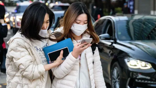 ソウルの梨花女子外国語高等学校の前で、毎年恒例の大学入試の受験に臨む生徒（右）。白いパフジャケットにマスク、リュックを背負い、携帯電話を見ている。彼女の隣では、青いポーチと携帯電話を持った年配の女性が、生徒の腕に右手を置いている。2022年11月17日撮影