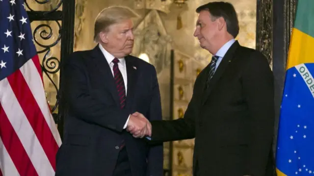 アメリカとブラジルの国旗を背に、共にダークスーツ姿で握手するトランプ氏とボルソナロ氏