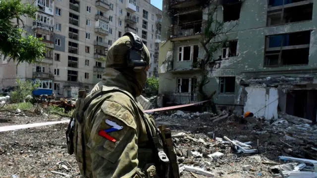 破壊されたウクライナ・セヴェロドネツクの住宅地に立つロシア兵。迷彩服を着て、ヘッドホンをしている。腕に、ロシア国旗の色で描かれた「Z」の標識がついている。