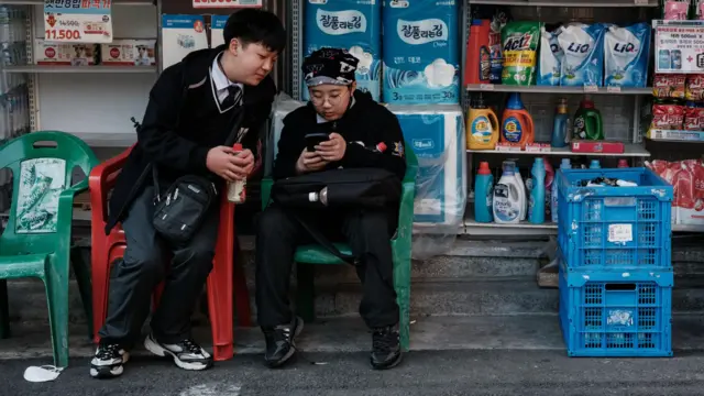 制服を着て眼鏡をかけた韓国の小学生が、コンビニエンスストアの外でプラスチックの椅子に座りながらスマホをスクロールしている。同じ制服を着た別の少年が、自分の椅子から身を乗り出して画面を見ている