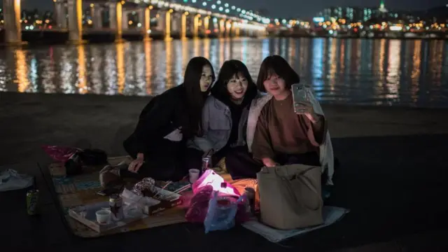 盤浦大橋の手前で自撮りのポーズをとりながら、笑顔で携帯電話を見つめる女性たち。草の上に毛布を敷き、暖かい服を着て座っている。前には食料容器とバッグがある。背後には湖とソウルの街並みが見える。2016年10月13日撮影