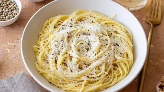 木のテーブルの上の白い皿に茹でたスパゲッティが盛られ、上に粉チーズと黒コショウがかかっている。周囲にはフォークやナプキン、水の入ったグラスなどが置いてある