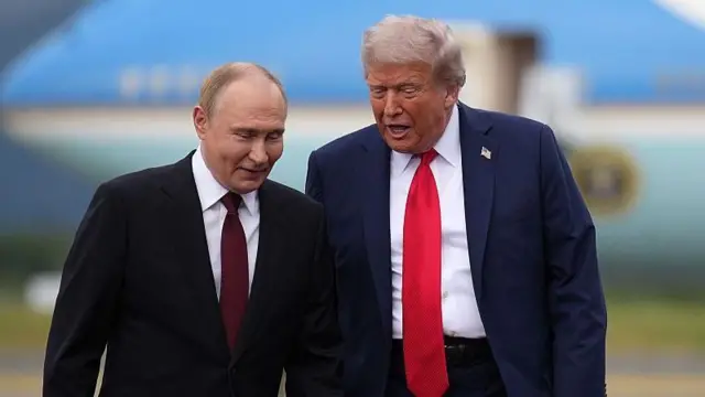 ダークスーツ姿の両首脳が横並びで立っている。トランプ氏が話をし、プーチン氏はほほえんでいる。背後には米大統領専用機が写っている