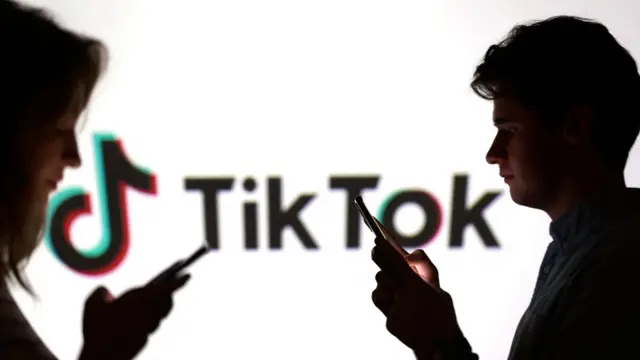 TikTokのロゴが映された大型モニターの前で、男女が向かい合い、それぞれスマートフォンを見ている。2人ともモニターからの逆光で影になっており、表情などは見えない