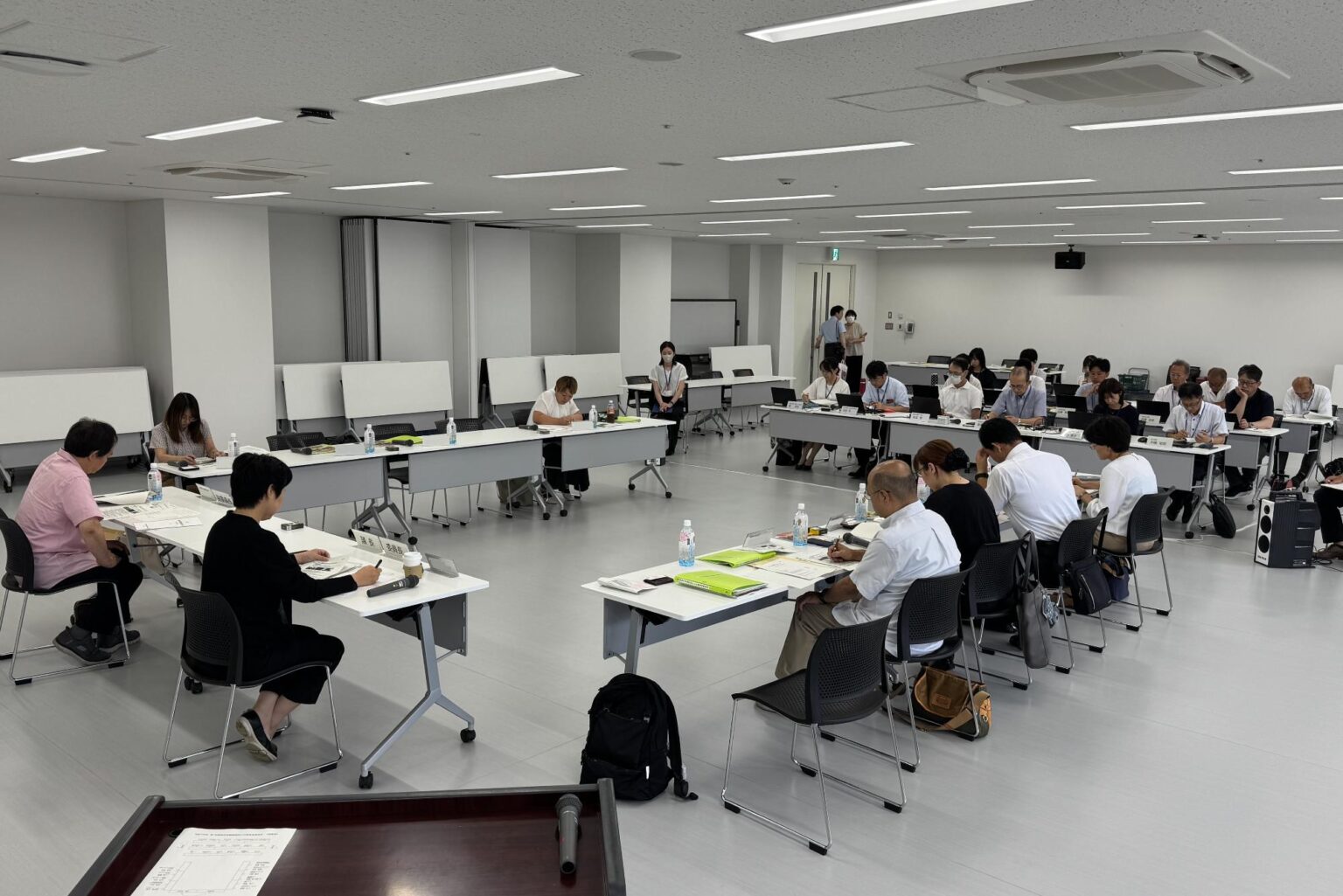 会議室にコの字型に並んだ会議机とその正面に横に数列並んだ会議机の席に出席者たちが座っている委員会の様子の写真