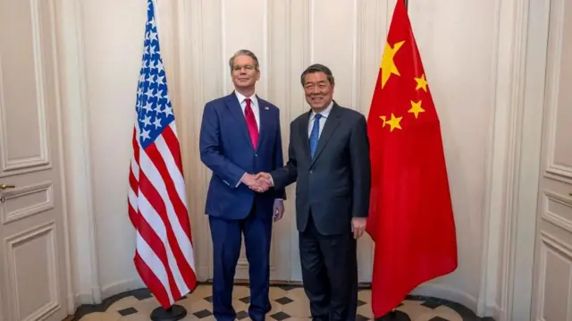 男性2人が、アメリカ国旗と中国国旗の間に立って握手を交わし、報道陣に笑顔を向けている