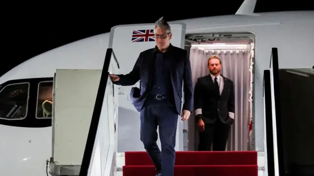 飛行機のタラップを下りるキアー・スターマー氏。ダークブルーのスーツに黒のシャツ。灰色の短髪で黒い眼鏡をかけている。開いたドアにはイギリス国旗が描かれている。ドアの奥には機内の客室乗務員が立っているのが見える。
