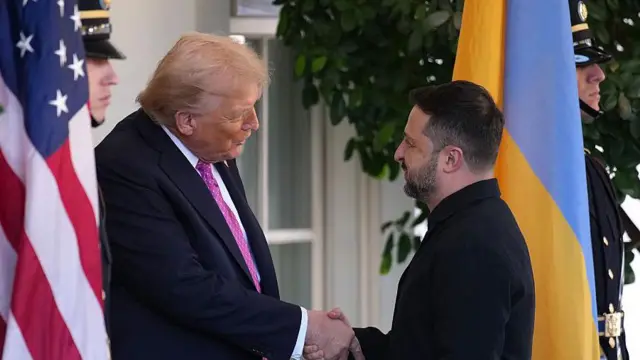 黒いジャケットを着たウクライナのゼレンスキー大統領を、ダークスーツに赤系のネクタイを着けたトランプ米大統領がホワイトハウスで出迎え、握手を交わしている。2人の両側にはアメリカとウクライナの国旗が置かれている