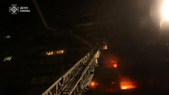 夜間に火災が発生し窓から炎が見えている高層ビルに、消防車のはしごが上層階に伸ばされているのを、下から撮った写真