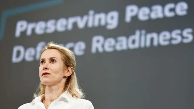 白いブラウスを着た金髪のカヤ・カラス氏が、「平和と防衛体制の維持」というスローガンが書かれたグレーの背景を前に立ち、前方を見ている