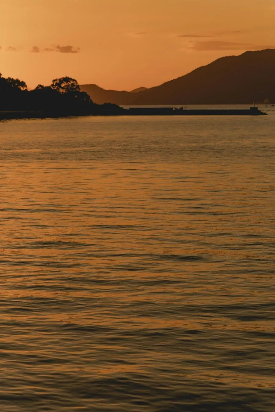 夕焼けに染まる海と山の風景。水面には夕日の光が反射し、穏やかな波が広がっている。