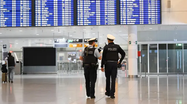 空港の発着便を表示する電光掲示板の下を警官2人が並んで歩いている。背中に「POLIZEI（ドイツ語で警察）」と書かれたベストを着ている。遠くにはスーツケースを引く男性の姿も見えるが、人はまばら