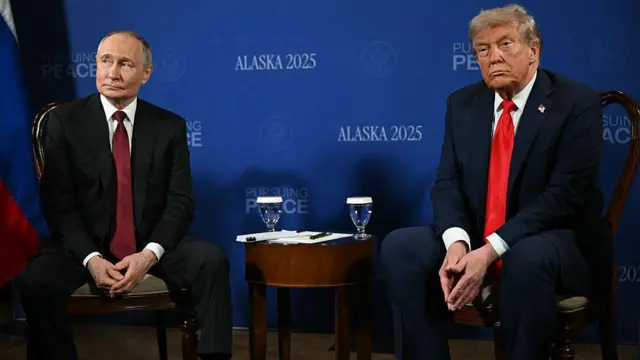 8月の首脳会談で隣同士に座るトランプ氏とプーチン氏。互いに別の方向を見ている。2人の間には丸井コービーテーブルが置かれ、メモとペン、水の入ったグラスが置かれている。背後には「ALASKA2025」と書かれた青いパネルが置かれている