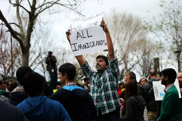 公園で「WE ARE ALL IMMIGRANTS（私たちは全員移民だ）」と書かれた紙を掲げるインド系の男性。周囲にもプラカードを持った人々が集まっている