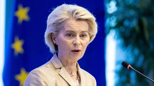 ベージュのブレザーを着た、白髪のフォン・デア・ライエン欧州委員長が、マイクに向かって話をしている。背後には、青地に金色の星が並ぶEUの旗がある
