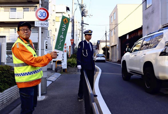 制限速度が引き下げられた小学校前で通行車両に注意を呼びかける下京署員（中央）とボランティア（２日、京都市下京区で）