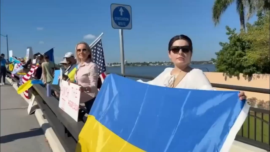 マール・ア・ラーゴへの橋に並ぶアメリカ人がウクライナの国旗を振り、ウクライナとトランプ大統領との会談のため向かうゼレンスキー大統領への支持を表明