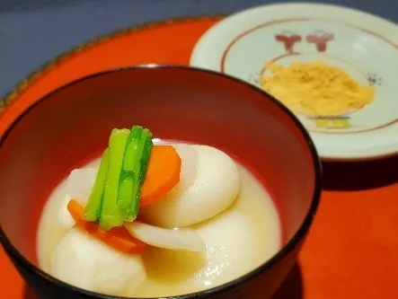 お椀に入ったお雑煮の写真。具材には、餅、野菜、だし汁が含まれている。奥には小皿が写っており、何かが盛られている。
