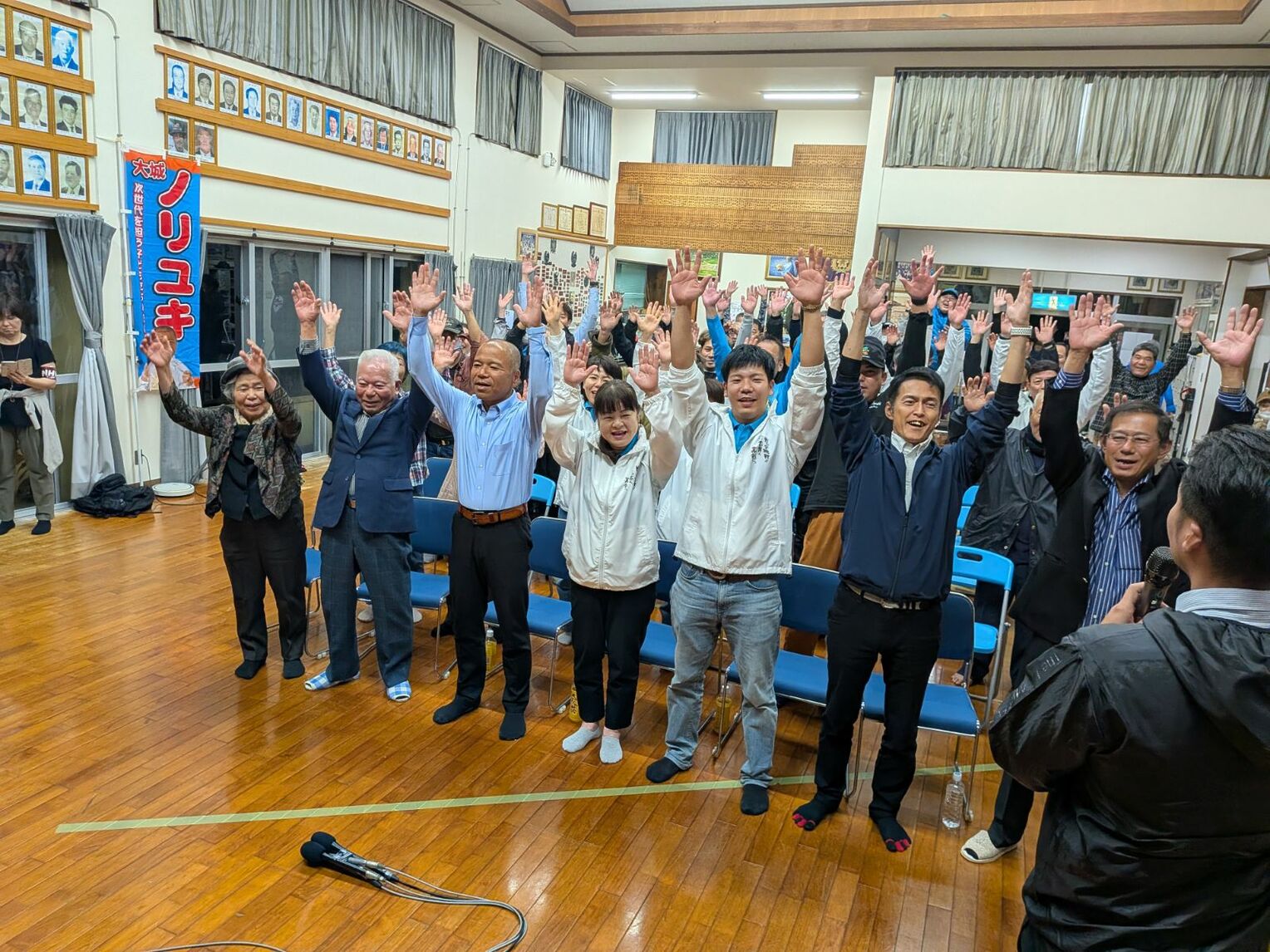 当選確実の一報を受けて、万歳する大城憲幸さん(左から3人目)=21日午後10時過ぎ、南城市の大城集落センター