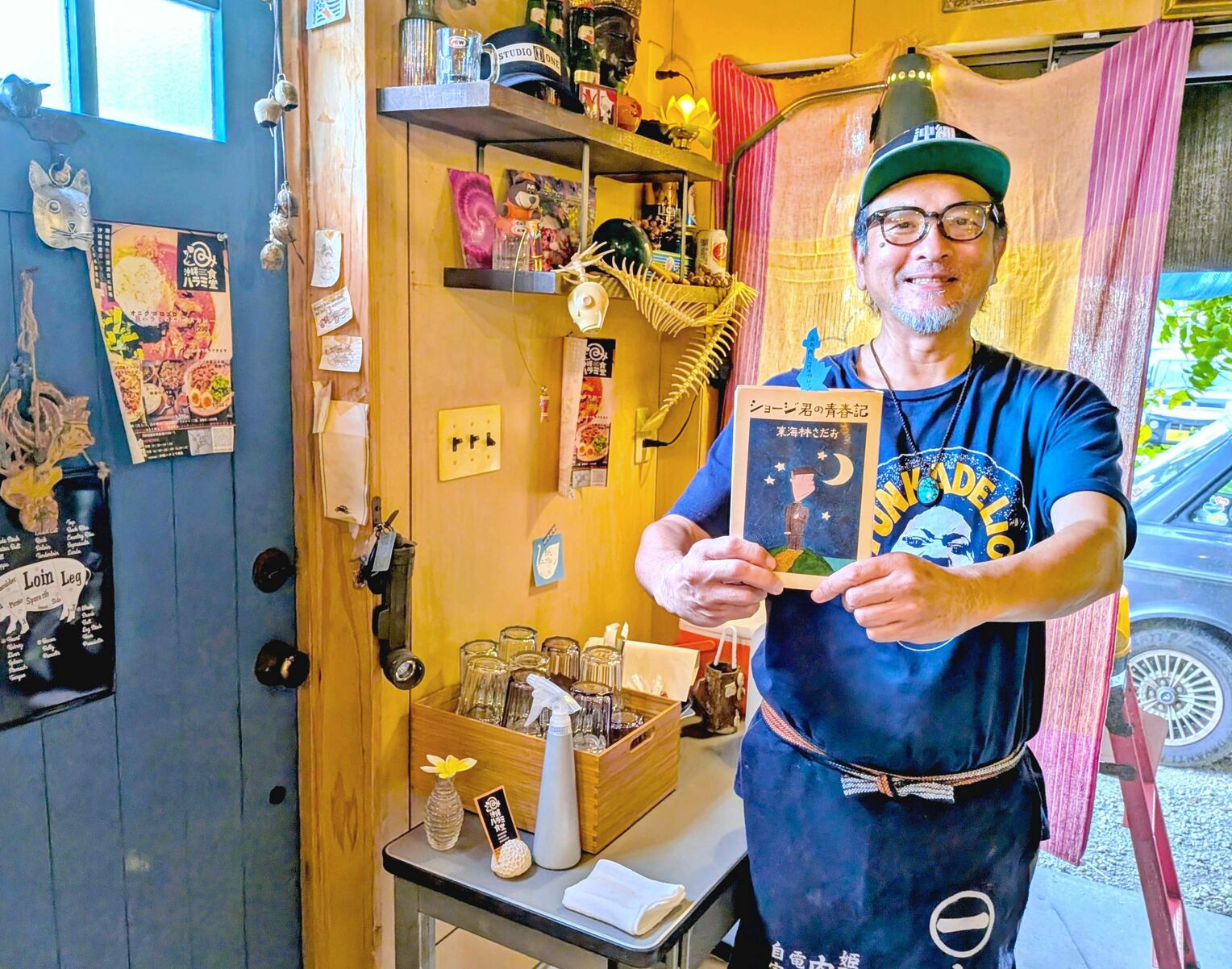 「ショージ君の青春記」を手にする秋満亜希さん。右奥は愛車のボルボ＝１１月２６日、南城市佐敷津波古