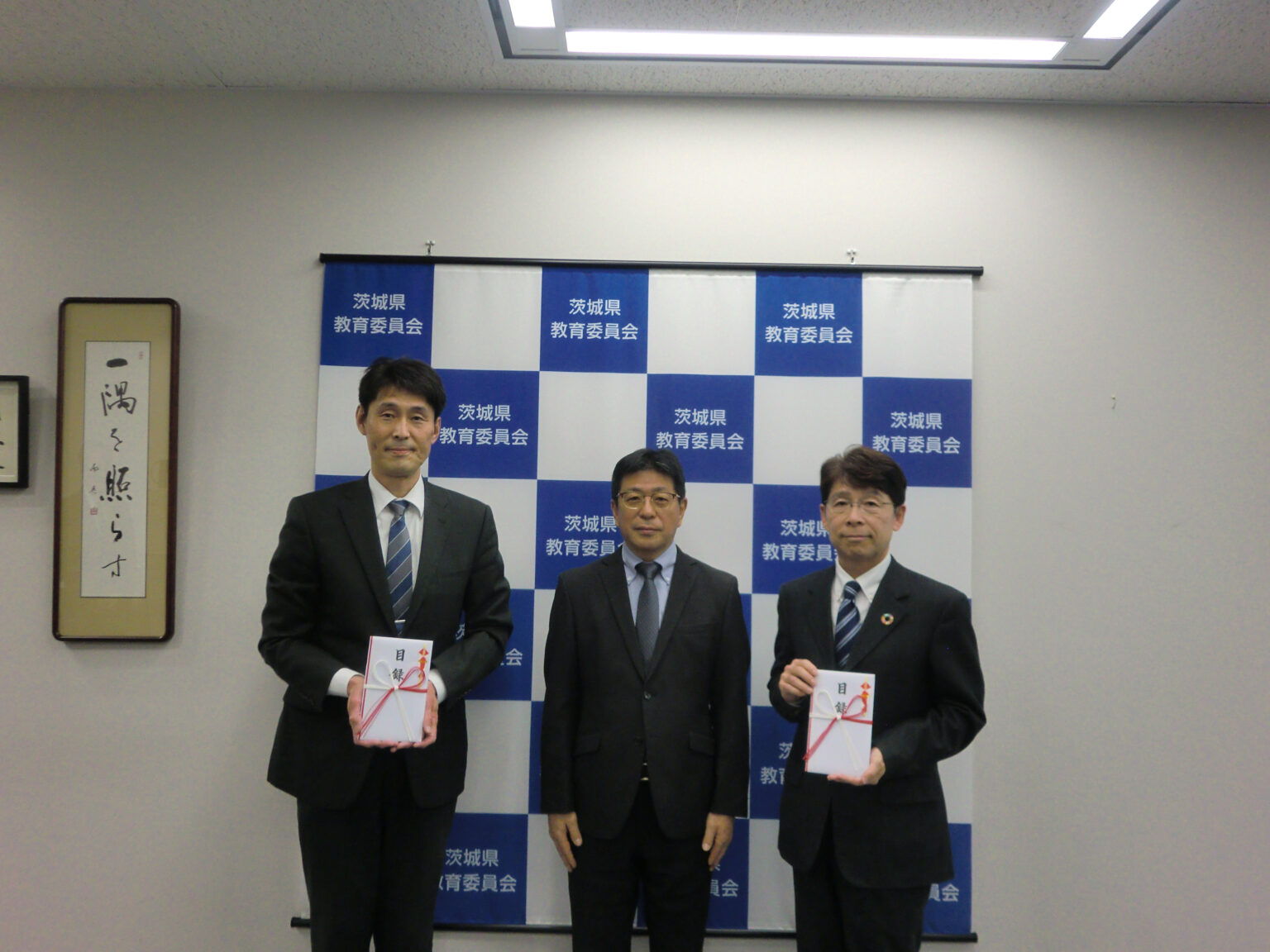 茨城県教育委員会のパネルを背景に男性が３人写っていて、２人が寄贈目録を持っている。