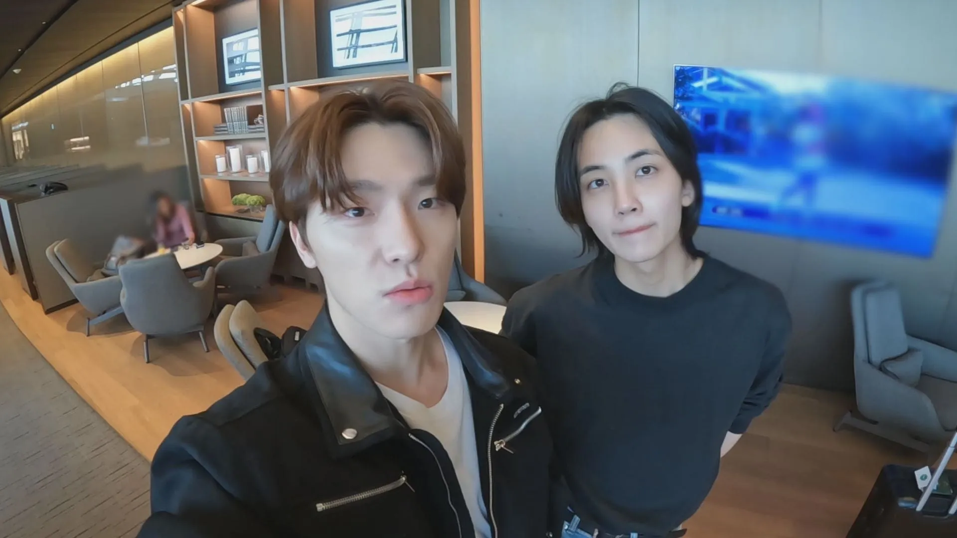 2人の男性がホテルのラウンジで自撮りしている様子。一人は黒い革のジャケットを着ており、もう一人は黒いセーターを着ています。背景にはテレビと座席があります。