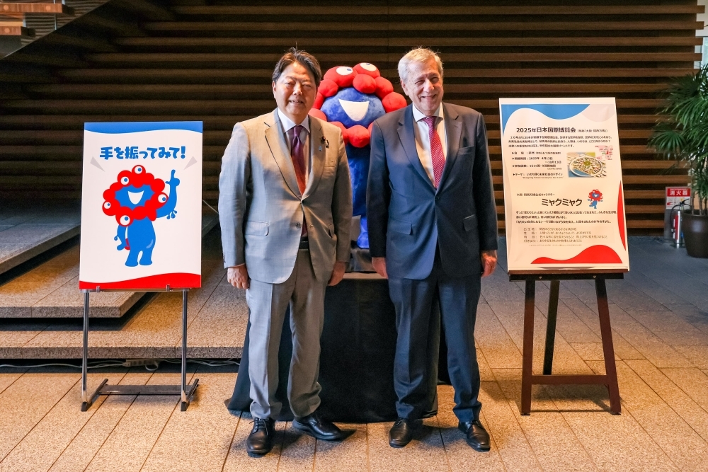 万博のキャラクター「みゃくみゃく」の人形を挟んで並ぶ林官房長官とクラベレン・チリ共和国外務大臣