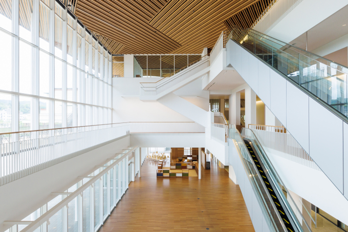 「ひろま」は3フロアの吹き抜けで、全面ガラス張りの窓で明るい空間。天井の木ルーバーは、鳥取砂丘の風紋や倉吉絣（かすり）の織模様を思わせる意匠で、洗練さの中に温もりを感じさせる。