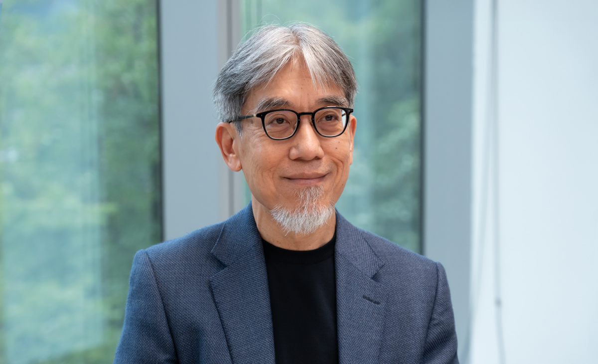 眼鏡をかけたモリスのバストショット写真。男性はグレーがかった髪をセンターで分け、整えられた白いあごひげをたくわえている。ネイビーのジャケットに黒いインナーを合わせ、穏やかな微笑みを浮かべている。