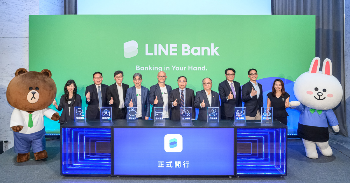LINE Bankのロゴと「Banking in Your Hand.」の文字が大きく表示された緑色の背景パネルの前で、複数の登壇者が横一列に並び、親指を立てるポーズをしている写真。中央にはスーツやジャケット姿の男女が並び、前方には青く発光する装飾が施されたステージ台が設置されている。ステージ中央にはLINE Bankのロゴと中国語で「正式開行」と表示されている。
