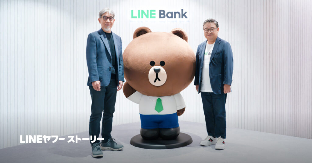 モリスとファンが、LINE Bankのロゴが掲げられた白い縦縞の背景の前で、大きなクマのキャラクターの像を挟んで立っている写真。中央には、白いシャツに緑のネクタイ、青いパンツを着用したクマのキャラクターが黒い台座の上に立っている。背景上部には「LINE Bank」のロゴが表示され、左下には「LINEヤフーストーリー」という文字が重ねて表示されている。