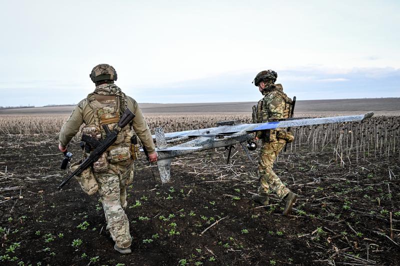 Two figures in camouflage gear walk on a brown field holding a flat grey large drone
