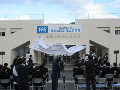 玄関入口前にパイプ椅子が複数台整列され参加者が着席しており関係者が紅白の紐を引っ張り白い布を下におろそうとしている除幕式の様子を撮影した写真