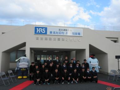 学生服を着た男女とスーツ姿の関係者が建物の入り口前に4列に並び両脇にサーモンくんとみやこちゃんも並び記念撮影をしている様子を撮影した写真