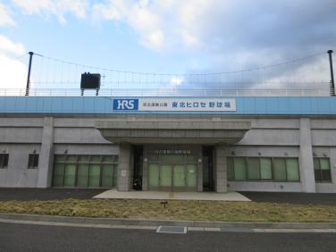 建物上部が水色で全体が灰色の外壁で「東北ヒロセ野球場」と書かれた看板が上部に設置されている玄関入口前の外観写真