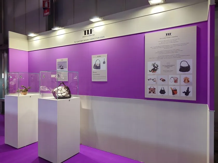 東京レザーフェアのTLFブース内に設置された革のデザインコンテスト歴代受賞作品展示コーナー。白いディスプレイ台上にバッグ2点をガラスケースに収め、紫の背景壁に過去受賞作一覧パネルを掲示した展示空間全景