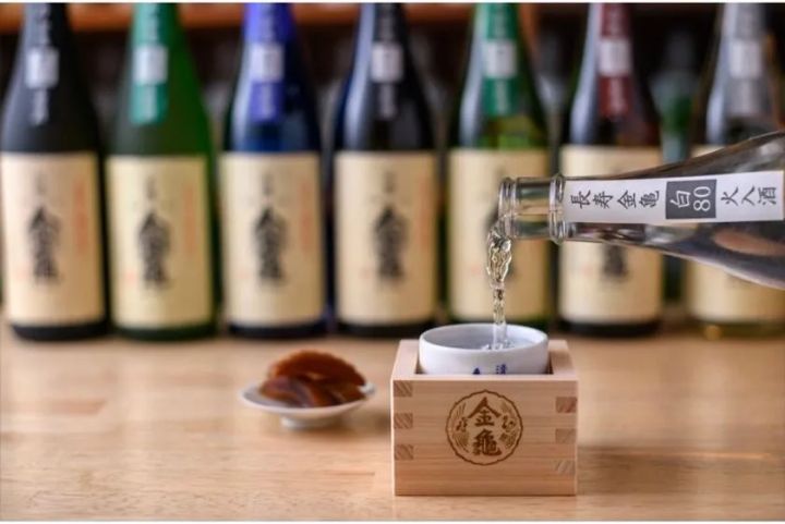 金亀ロゴ入り升に日本酒を注ぐ試飲シーン。背景に複数の金亀銘柄ボトルが並び、酒肴の小皿も添えられた酒蔵試飲体験の様子